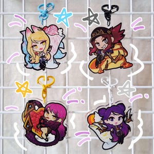 Peut inclure: Quatre breloques en acrylique représentant des personnages de dessins animés aux couleurs vives, avec des porte-clés. Les breloques incluent un personnage blond avec une glace, un personnage avec un chapeau "KDA", un personnage avec un cœur et un personnage avec une flamme.