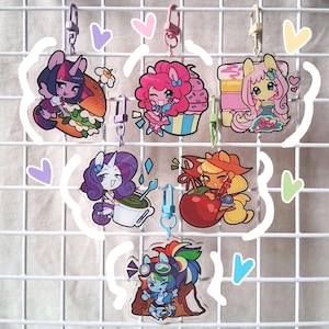 Puede incluir: Una colección de llaveros acrílicos coloridos con personajes de dibujos animados de ponis. Cada llavero tiene un diseño de personaje diferente, con varias poses y accesorios. Los llaveros están sujetos a cierres metálicos y se exhiben sobre un fondo de rejilla blanca.