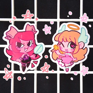 May include: Two cartoon stickers featuring cute characters. One is a pink devil with a mirror, the other is an angel with a halo and wings. Pink stars and dots surround the stickers on a black grid.
