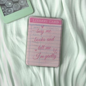 Puede incluir: Una funda de teléfono rosa con diseño de tarjeta de biblioteca con el texto "buy me books and tell me I'm pretty". La funda está sobre una tela verde claro, junto a un lector electrónico verde menta.