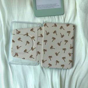 Peut inclure: Un étui clair pour liseuse électronique avec un motif de palmiers. L'étui est ouvert, révélant l'intérieur et un support en plastique transparent. La liseuse est vert clair. Le fond est un tissu vert clair.