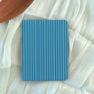 Könnte beinhalten: Eine blaue Tablet-Hülle mit einem vertikalen Streifenmuster in verschiedenen Blautönen. Die Hülle ist rechteckig mit abgerundeten Ecken und scheint aus einem glatten Material zu bestehen. Der Hintergrund ist ein weißer, strukturierter Stoff.