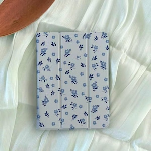 Pode incluir: Uma capa branca para tablet com um padrão floral azul. A capa apresenta um design repetido de pequenas flores e folhas azuis. A capa é retangular e tem uma capa dobrável.