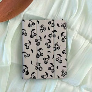 May include: A light-colored tablet case featuring a repeating pattern of black cherries with the number 8 in the center of each cherry. The case has a smooth, matte finish and a magnetic closure.