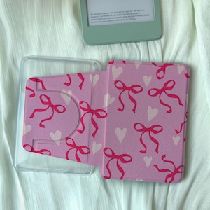 Puede incluir: Funda rosa para Kindle con un estampado de lazos rojos y corazones blancos. La funda está abierta, revelando un soporte de plástico transparente y un dispositivo Kindle verde menta en el fondo. La funda está diseñada para proteger el Kindle.