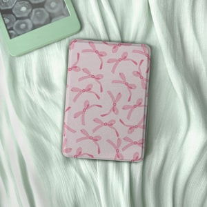 Puede incluir: Una funda rosa y blanca para lector de libros electrónicos con un patrón repetido de lazos rosas. La funda es rectangular y descansa sobre una tela verde claro texturizada. Un lector de libros electrónicos verde menta es parcialmente visible en la esquina superior izquierda.