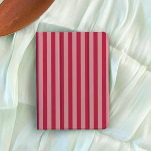 May include: A rectangular tablet case with a vertical striped pattern in shades of pink and red. The case is resting on a light green, textured fabric. The case appears to be closed.