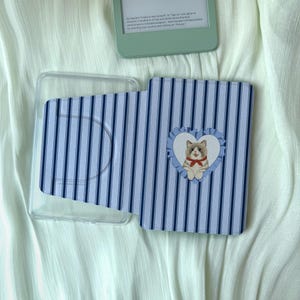以下が含まれることがあります： 青と白のストライプのKindleケースと透明なプラスチックホルダー。ケースには、赤い蝶ネクタイを着た漫画の猫のハート型のデザインが施されています。ミントグリーンのKindleが背景に見えます。