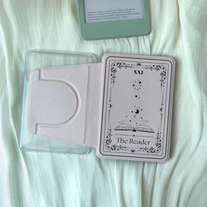 Op de afbeelding: Een lichtroze e-reader hoesje met een tarotkaart ontwerp. De kaart toont de tekst "The Reader" en een hemelse illustratie. Een doorzichtig plastic hoesje is zichtbaar naast het open hoesje, en een mintgroene e-reader staat op de achtergrond.
