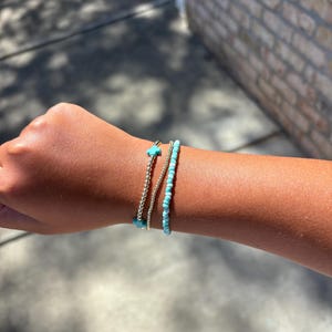 May include: Three bracelets on a wrist. One is a silver chain, another is a string of small turquoise beads, and the third is a silver chain with a turquoise cross charm. The bracelets are stacked together.