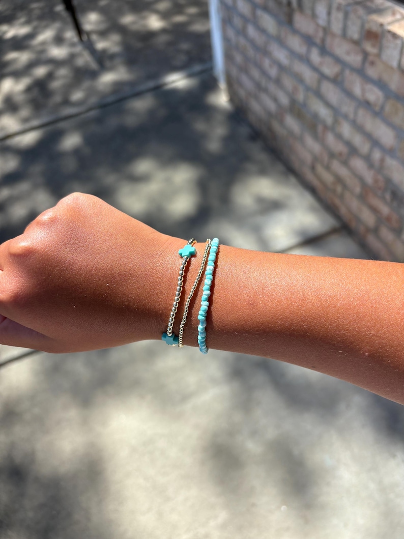 May include: Three bracelets on a wrist. One is a silver chain, another is a string of small turquoise beads, and the third is a silver chain with a turquoise cross charm. The bracelets are stacked together.