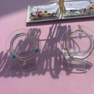 May include: Gold beaded bracelets with turquoise cross charms are displayed on clear stands. The bracelets are presented on a pink surface, with additional jewelry and beads visible in the background.
