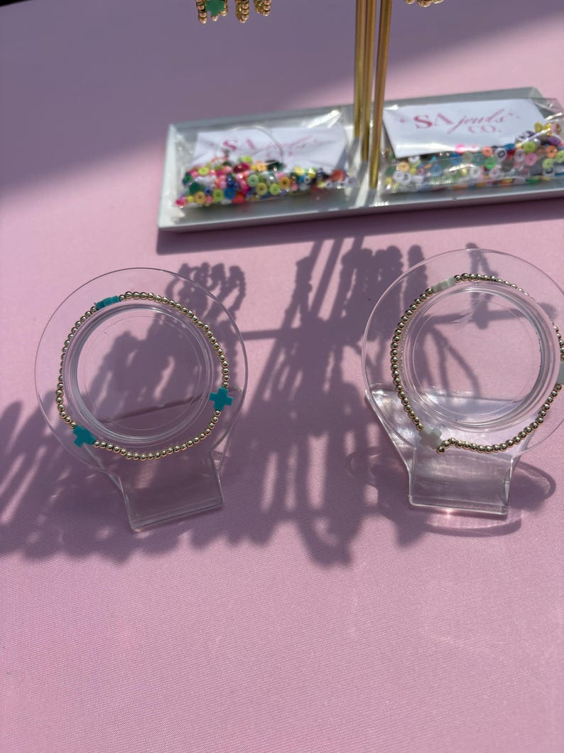 May include: Gold beaded bracelets with turquoise cross charms are displayed on clear stands. The bracelets are presented on a pink surface, with additional jewelry and beads visible in the background.