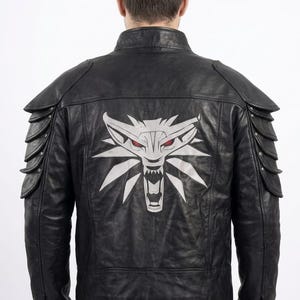 Puede incluir: Una chaqueta de cuero negro con cuello alzado y detalles de hombreras superpuestas. La espalda muestra un gran emblema de cabeza de lobo plateado con llamativos ojos rojos. La chaqueta tiene mangas largas con puños abotonados y corte entallado.