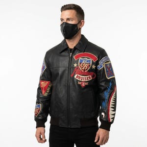 Könnte beinhalten: Eine schwarze Leder-Bomberjacke mit Frontreißverschluss, Kragen und gerippten Bündchen. Die Jacke ist mit verschiedenen gestickten Aufnähern verziert, darunter der Schriftzug American Bruiser, Honor & Duty und eine Haifischmaul-Grafik am Ärmel.