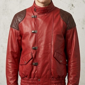 May include: A red leather jacket featuring dark brown quilted shoulder panels, a high collar with a buckle strap, and a front closure with four metal buckles. The jacket includes two side pockets with flaps and a ribbed hem at the waist.
