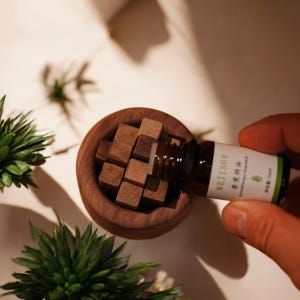 Peut inclure: Un bol en bois rempli de petits blocs carrés bruns. Une bouteille en verre brun étiquetée "MEJISOO" verse du liquide dans le bol. La bouteille a une étiquette blanche avec des accents verts. L'image a une esthétique naturelle, de type spa.