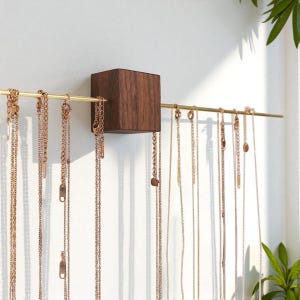 Può includere: Un organizer di gioielli da parete con un cubo di legno scuro e una barra di metallo color oro. Varie collane in tonalità oro e rame sono appese alla barra. Il design è minimalista e moderno, adatto per esporre collane.