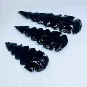 May include: Three black obsidian arrowheads with serrated edges are shown against a white backdrop. The arrowheads have a glossy, reflective surface, and vary slightly in size. The overall aesthetic is reminiscent of ancient tools or artifacts.