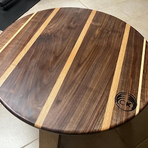 May include: A round wooden table with a dark brown surface and light brown stripes. The table has a logo on the right side. The table is supported by a light-colored wooden base. The table is approximately 30 inches in diameter.