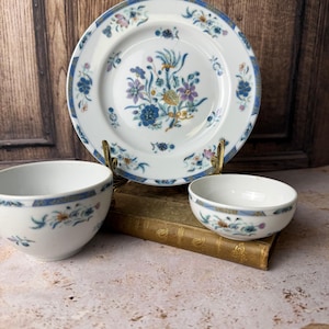 Puede incluir: Un plato de porcelana blanca y dos cuencos a juego con un estampado floral en azul, morado y amarillo. El plato se exhibe en un soporte dorado, mientras que los cuencos están colocados delante. El conjunto incluye un cuenco grande y uno pequeño, decorados con delicados motivos botánicos y bordes azules.