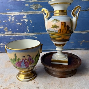Puede incluir: Una urna de porcelana blanca con bordes dorados y un paisaje pintado con un edificio y árboles, junto a una taza de porcelana blanca con bordes dorados y una escena pintada de dos figuras con ropa de época, ambas con dos asas laterales.