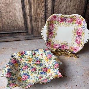 May include: Two decorative porcelain trays with floral patterns. One tray is square with a scalloped edge and a colorful floral design. The other tray is rectangular with a pink rose border and gold trim.