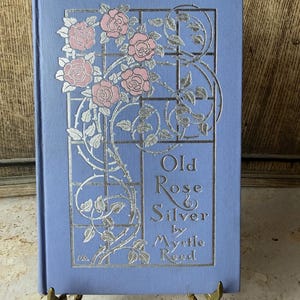 以下が含まれることがあります： Myrtle Reedの「Old Rose & Silver」というタイトルのヴィンテージ本。本は、銀色の花の模様とピンクのバラが付いた水色の表紙です。本は金色のスタンドに展示されています。