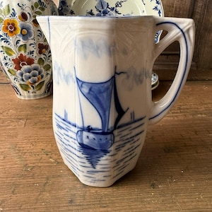 May include: White ceramic pitcher with a blue sailboat design. The pitcher has a rectangular shape with a curved handle. Other ceramic pieces with floral and blue patterns are in the background.