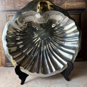 May include: A silver-toned, shell-shaped serving dish with a fluted design. The dish has a gold-colored accent and is supported by a black stand. The dish is likely made of metal and is designed for serving food or displaying decorative items.