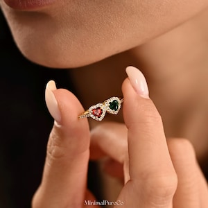 Könnte beinhalten: Ein goldener Ring mit zwei herzförmigen Edelsteinen, einem roten und einem grünen, die jeweils von kleinen, klaren Steinen umgeben sind. Der Ring wird von Fingern gehalten, was das filigrane Design hervorhebt. Der Ring ist ein Schmuckstück.