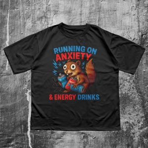 Könnte beinhalten: Schwarzes T-Shirt mit dem Text "RUNNING ON ANXIETY & ENERGY DRINKS". Die Grafik zeigt ein Cartoon-Eichhörnchen, das zwei Energy-Drink-Dosen hält. Das Eichhörnchen ist braun mit rotem Schwanz und hat große Augen.