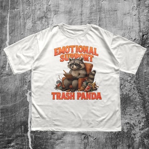 Könnte beinhalten: Weißes T-Shirt mit einem Waschbären mit Brille, der in einem Stuhl sitzt und eine Schüssel hält. Der orangefarbene Text lautet "EMOTIONAL SUPPORT TRASH PANDA". Der Waschbär ist von Müll umgeben.