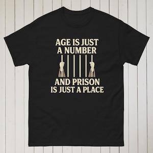 Puede incluir: Camiseta negra con el texto blanco "AGE IS JUST A NUMBER AND PRISON IS JUST A PLACE". El diseño incluye manos agarrando barrotes de prisión. La camiseta se muestra sobre un fondo de madera clara.