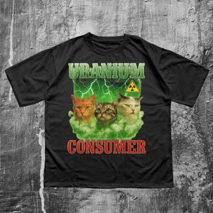 以下が含まれることがあります： 黒いTシャツ。緑色の雲の中に3匹の猫が描かれ、「URANIUM CONSUMER」の文字が緑と赤で書かれています。黄色の放射線マークも付いています。デザインはヴィンテージで、少し使い古されたような外観です。