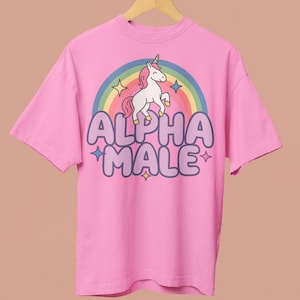 Könnte beinhalten: Ein pinkfarbenes T-Shirt mit einem Einhorn-Regenbogen-Motiv und dem Text "ALPHA MALE" in Lila. Das T-Shirt hängt an einem Holzkleiderbügel. Der Hintergrund ist rosa.