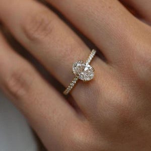 May include: A gold engagement ring featuring an oval-cut diamond as the centrepiece. The band is embellished with smaller diamonds, adding to the ring's sparkle. The ring is displayed on a finger, highlighting its design and elegance.