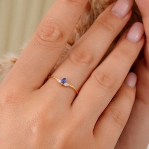 Peut inclure: Une délicate bague en or ornée d'une pierre saphir taille marquise flanquée de deux petites pierres blanches en forme de poire. La bague est sertie sur un fin anneau en or. La bague est présentée sur une main aux ongles manucurés.