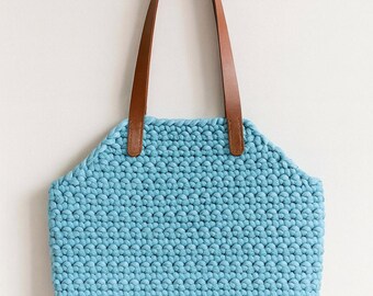 Könnte beinhalten: Hellblaue Häkeltasche mit braunen Ledergriffen. Die Tasche hat ein strukturiertes, gewebtes Aussehen und eine rechteckige Form mit gebogenen Seiten. Die Griffe sind oben an der Tasche befestigt.
