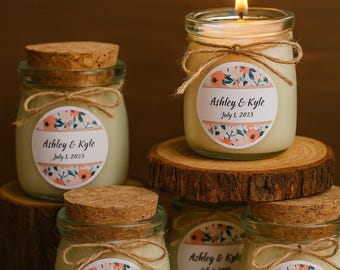 May include: A collection of small glass jar candles with cork lids and twine detailing. Each candle features a label reading "Ashley & Kyle July 1, 2023" with floral patterns. One candle is lit, the others unlit. Candles are displayed on wooden coasters.