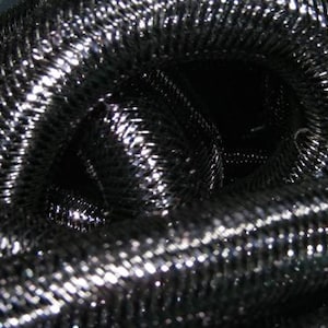 Puede incluir: Una manguera metálica flexible y enrollada con un exterior de malla de alambre plateado y negro estrechamente tejido. La superficie muestra un patrón diagonal repetitivo creado por los hilos entrelazados, con reflejos brillantes en el metal.