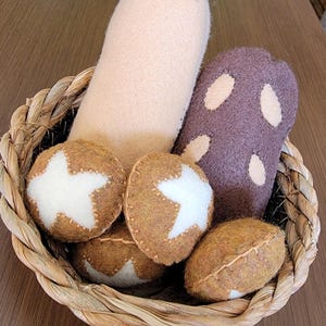 Puede incluir: Una cesta tejida llena de juguetes de fieltro hechos a mano. La cesta contiene una forma cilíndrica beige claro, una forma morada con manchas blancas y varios objetos marrones en forma de huevo con diseños de estrellas blancas. La cesta está sobre una superficie de madera.