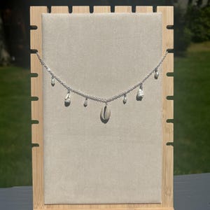 Puede incluir: Un collar de plata con un colgante de concha de cauri y varios pequeños abalorios de concha. El collar se exhibe en un soporte cubierto de tela beige con un marco de madera natural.