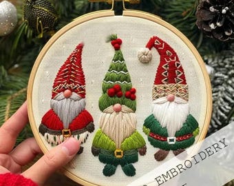 Könnte beinhalten: Gestickte Weihnachts-Wichtel-Hoop-Art. Drei Wichtel mit roten, grünen und weißen Details sind abgebildet. Die Wichtel haben lange weiße Bärte und tragen festliche Hüte und Outfits. Der Text "HAND EMBROIDERY KIT" ist sichtbar.