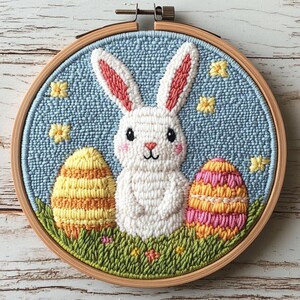 May include: A handmade Easter-themed embroidery hoop featuring a white bunny with pink-lined ears, two decorated eggs, and yellow flowers on a light blue background. The design is set in a wooden hoop, perfect for spring decor.