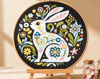May include: A round embroidery hoop featuring a white rabbit design with colorful floral accents on a black background. The rabbit is surrounded by flowers in shades of blue, yellow, pink, and green. The hoop is displayed on a small wooden easel.