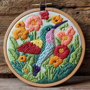 May include: A colorful embroidered hummingbird and floral design in a wooden hoop. The hummingbird features shades of green, blue, pink, yellow, and red. The flowers are orange, red, and pink, surrounded by green leaves. The embroidery is on a cream-colored fabric.