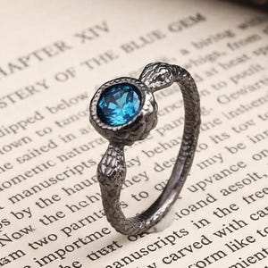 Puede incluir: Un anillo plateado con una gema azul, colocado sobre una página de libro. El anillo presenta un diseño de serpiente. El texto del libro incluye el título del capítulo "El Misterio de la Gema Azul".