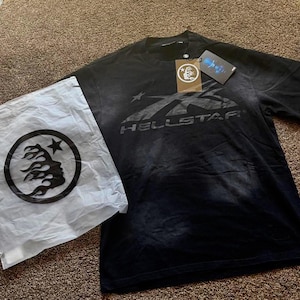 Könnte beinhalten: Schwarzes T-Shirt mit dem Wort "HELLSTAR" in einem verblassten grauen Design. Das Shirt hat einen Rundhalsausschnitt und kurze Ärmel. Eine weiße Tasche mit schwarzem Logo und ein Etikett liegen neben dem Shirt.