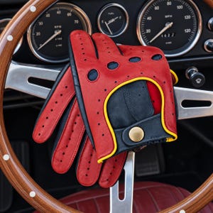 May include: A pair of red and black leather driving gloves featuring perforated detailing, a yellow trim along the wrist opening, and a brass-coloured snap button closure. The gloves are positioned over a wooden steering wheel with a metal centre.